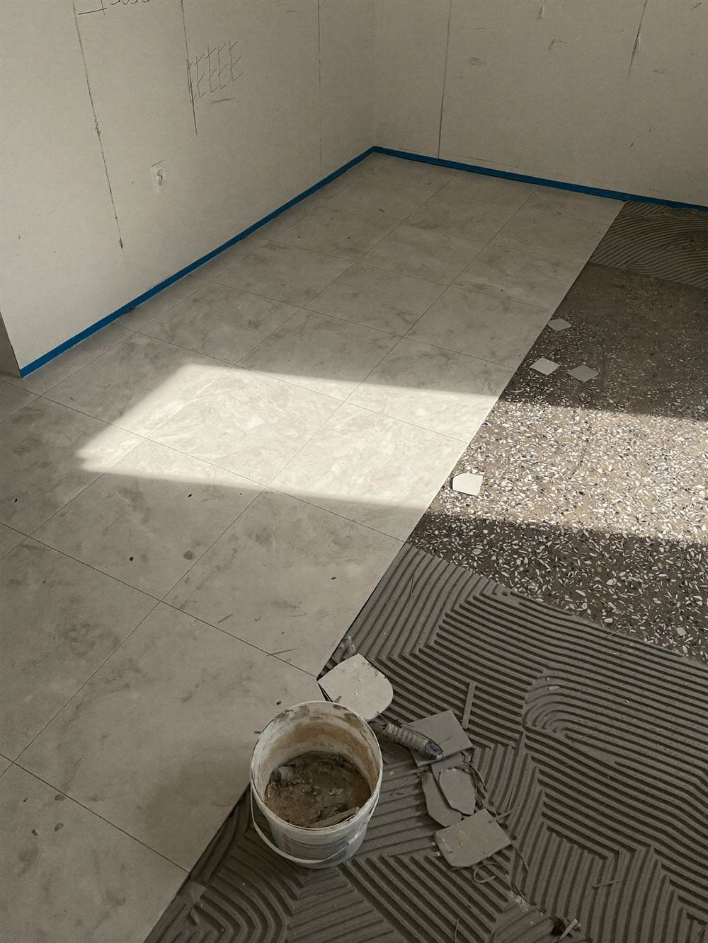 A floor half laid with pale ceramic tiles, the rest still bare terrazzo, with combed adhesive, tile spacers and offcuts.