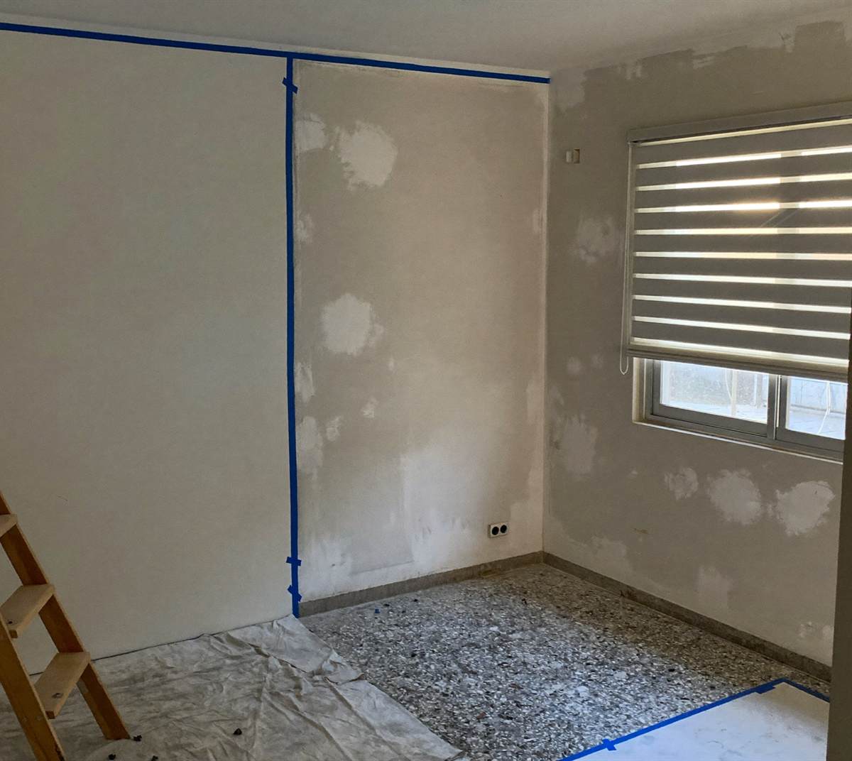 A room being repainted, with masking tape down the corner and along the floor, filler patches on the plaster and a dust sheet on the floor.